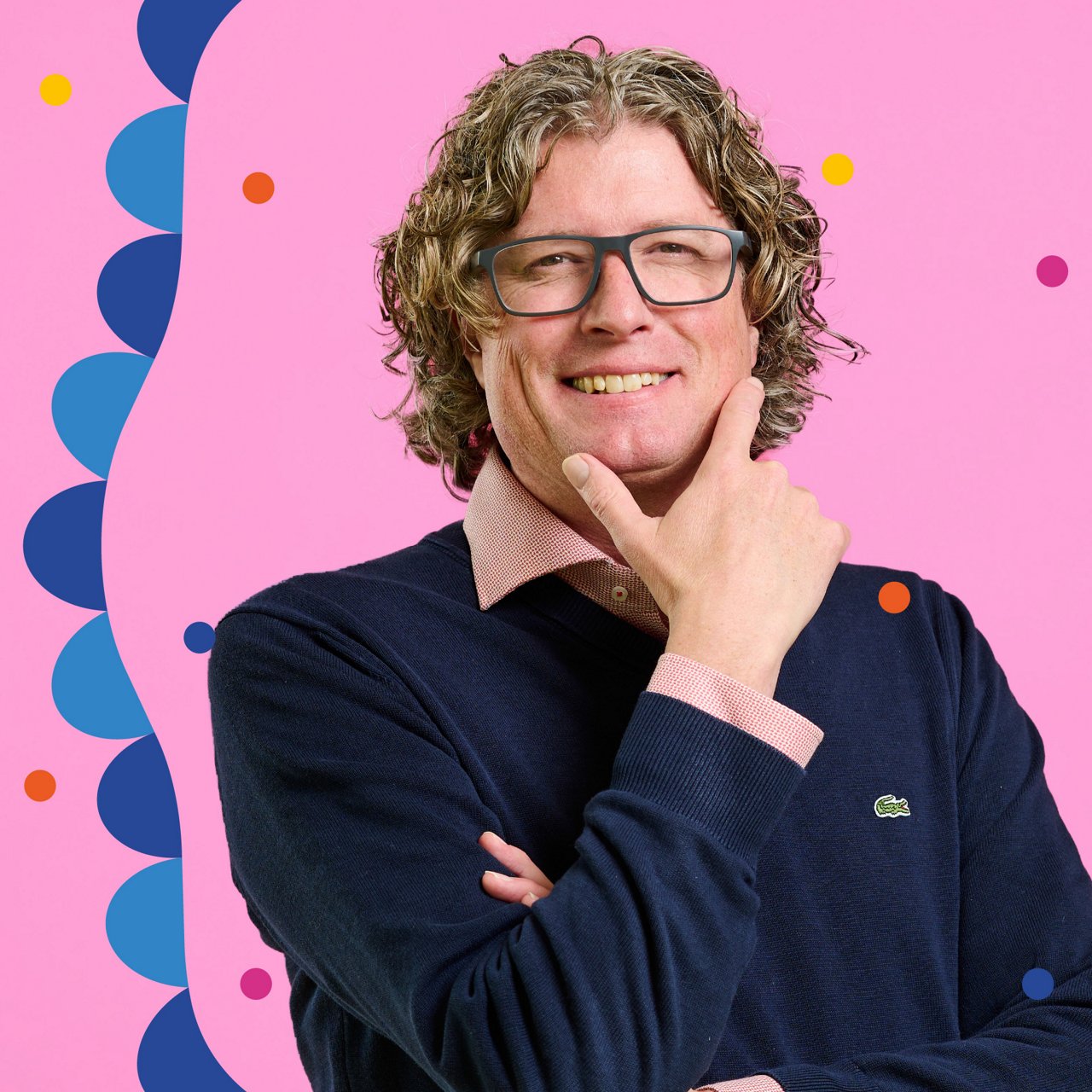 Portrait of a man with medium-length curly hair and glasses against a pink background. He wears a dark blue sweater over a pink shirt. His arms are crossed, and one hand touches his chin while he smiles warmly at the camera