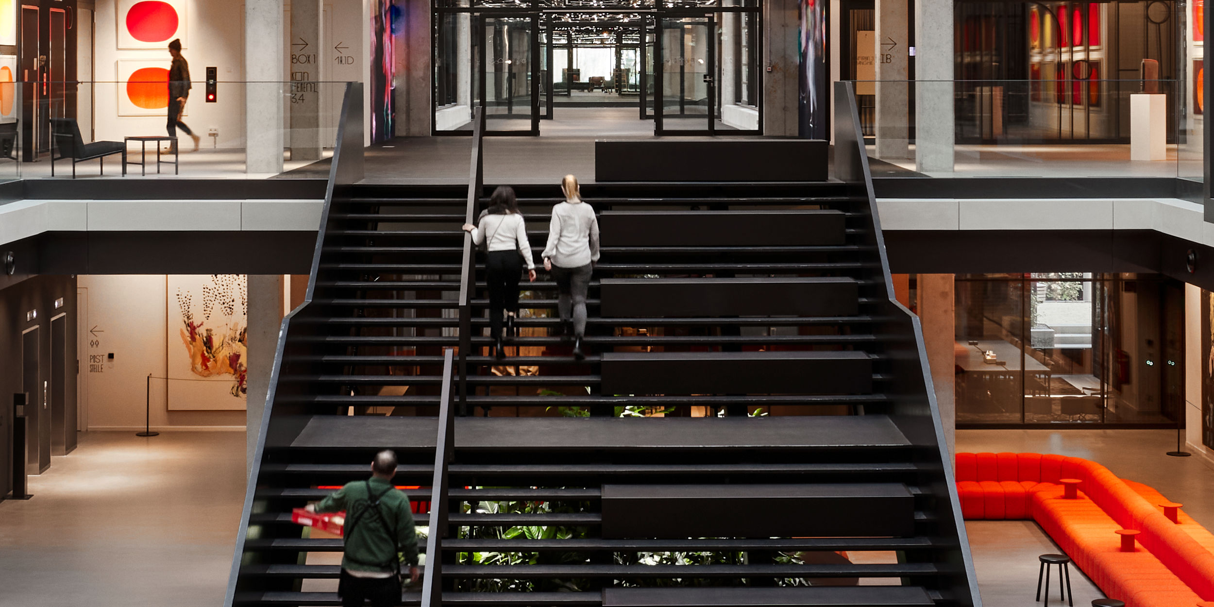 The foyer of the House of Communication Munich. In the centre, the open staircase from the ground floor to the first floor on which two people are walking.