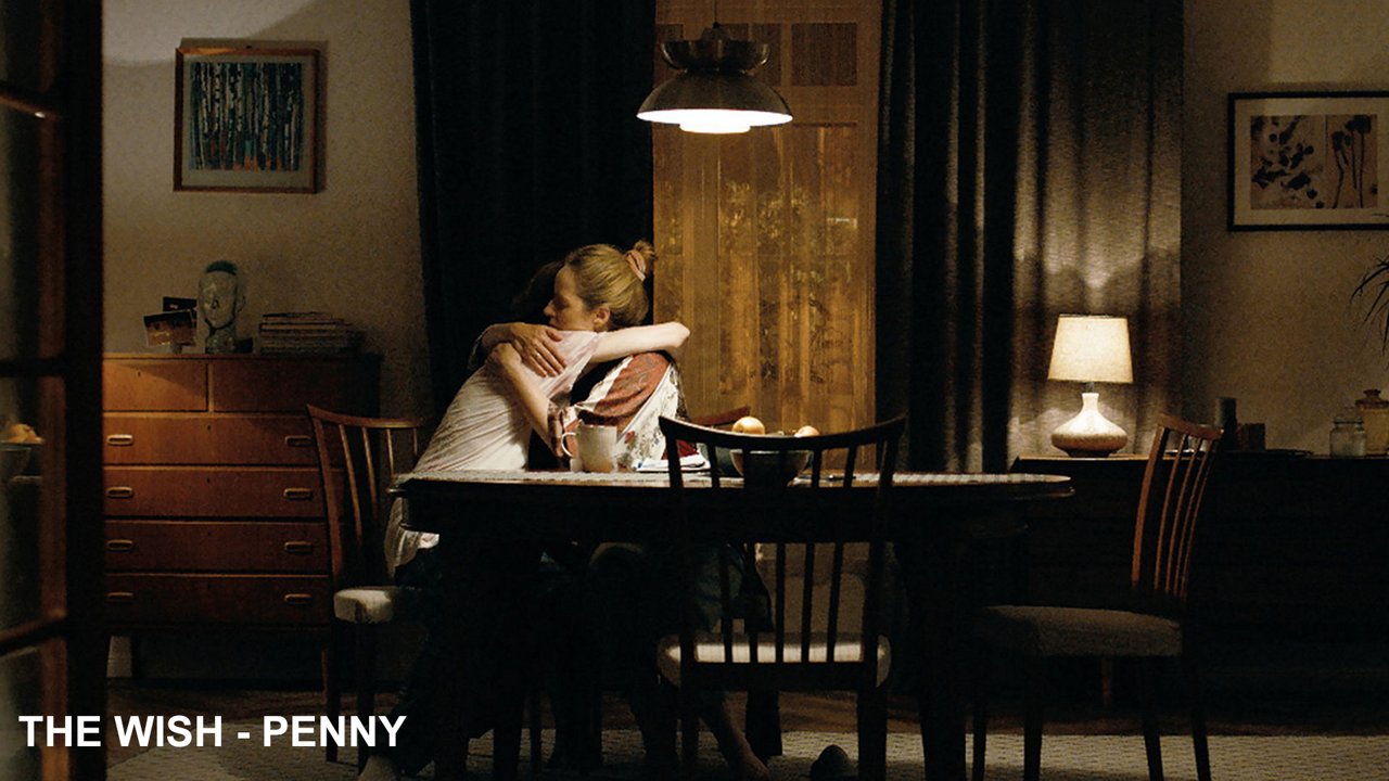 Two people sitting in an poorly lit apartment at a table hugging each other